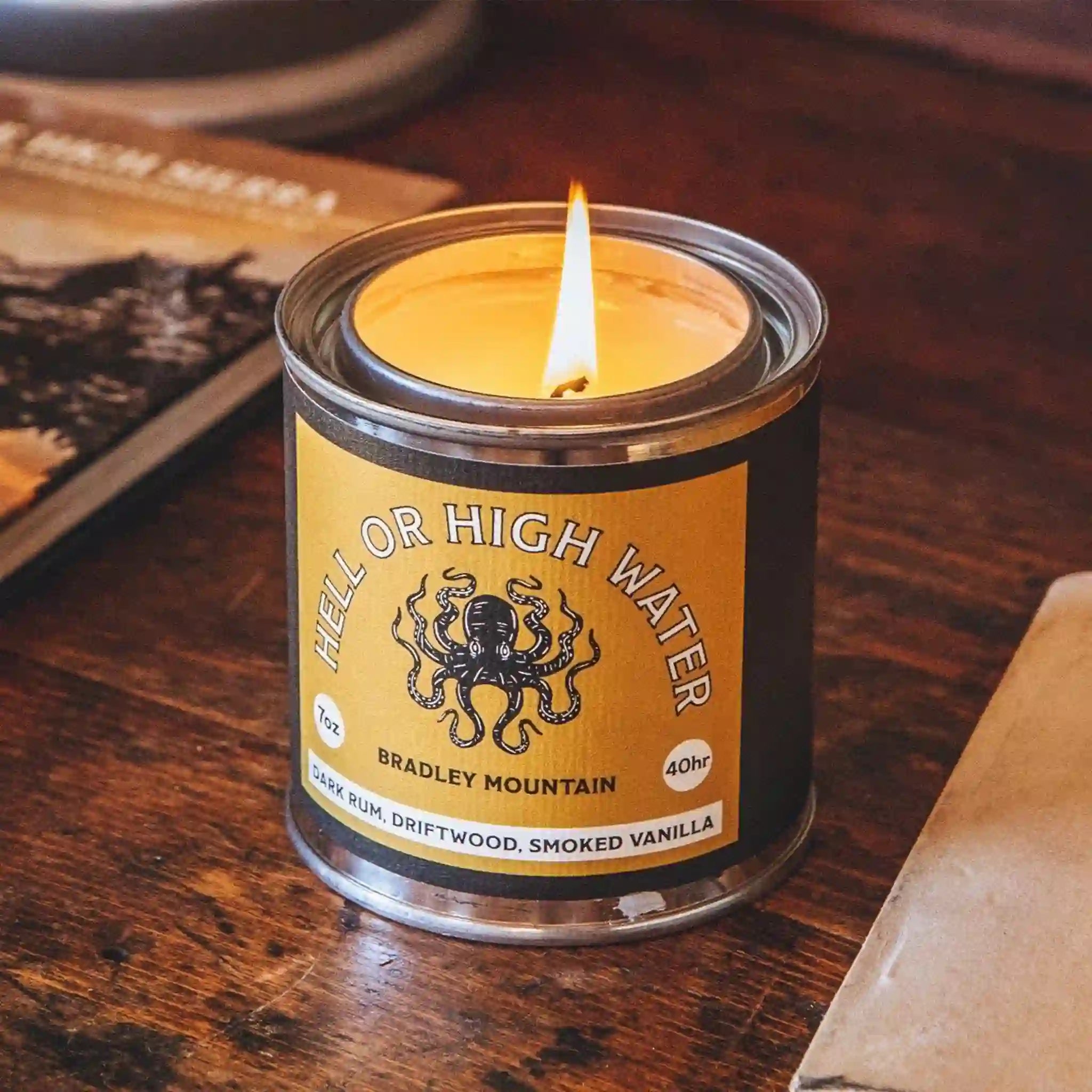 On a wood background is a tin candle with a yellow label featuring a octopus graphic and text arched above it that reads, &quot;Hell or High Water Bradley Mountain&quot;. 