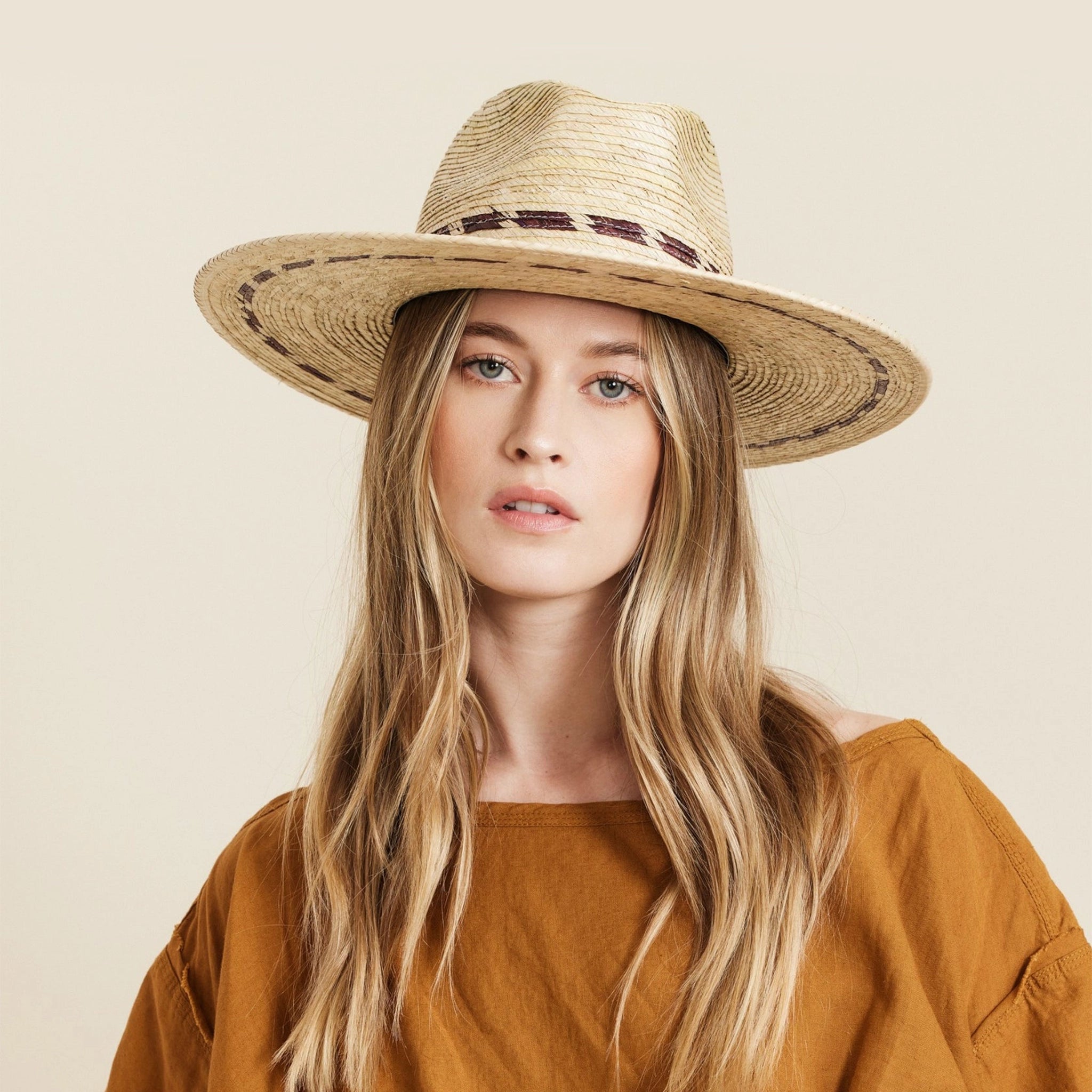 On a neutral background is a model wearing a tan woven sun hat with a wide brim and brown stitching details around the bottom of the brim as well as around the base of the hat. 