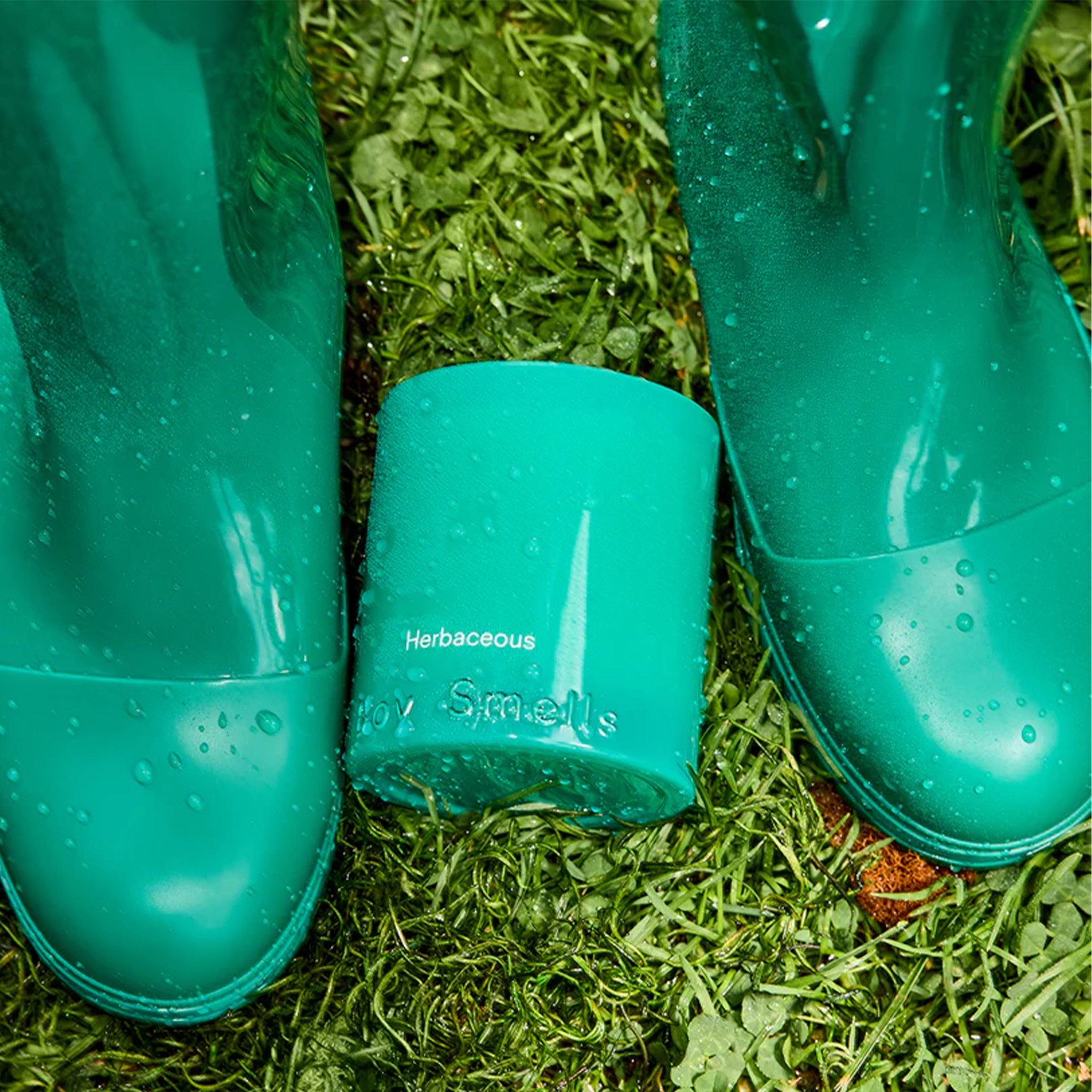 A teal glass candle with text that reads, 'Herbaceous Boy Smells'. 