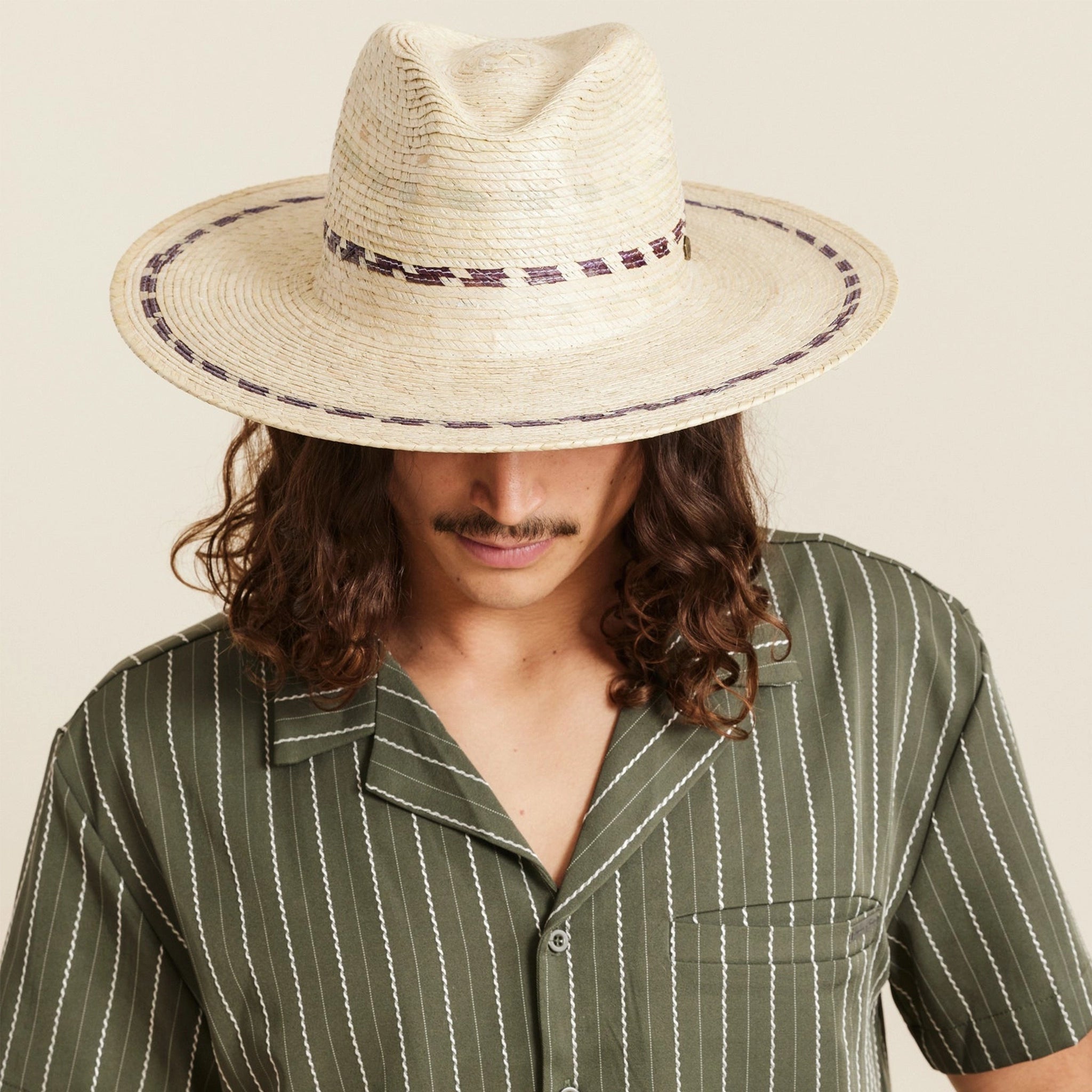On a neutral background is a model wearing a tan woven sun hat with a wide brim and brown stitching details around the bottom of the brim as well as around the base of the hat.
