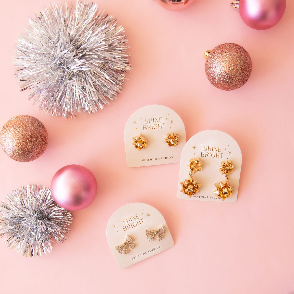 Three of Sunshine Studios bow shaped earrings on the packaging and next to ornaments and tinsel. 