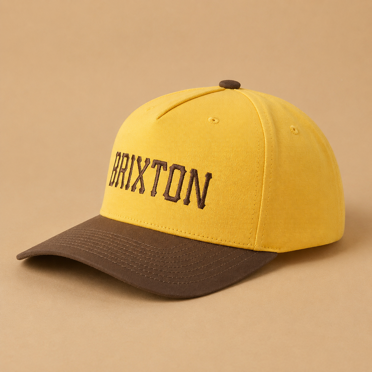A yellow and brown baseball cap with 'BRIXTON' embroidered in brown on the front, set against a solid beige background.