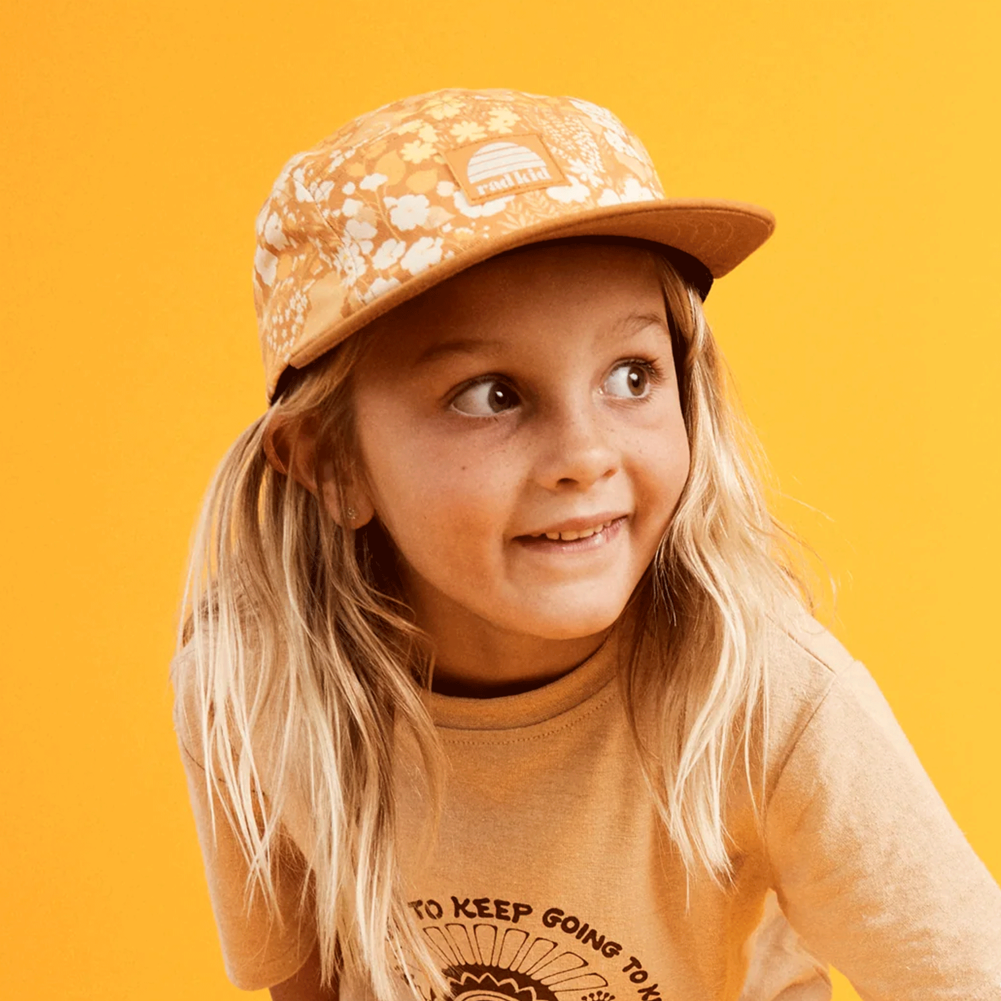 On a yellow background is a children&#39;s model wearing a yellow and brown floral print hat with a square label on the front that reads, &quot;rad kid&quot;.