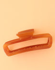 On a peachy background is an amber colored rectangle claw clip with slightly rounded edges.