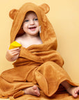On a yellow background is a caramel orange colored bath towel for kids with a hood and bear ears on the hood. 