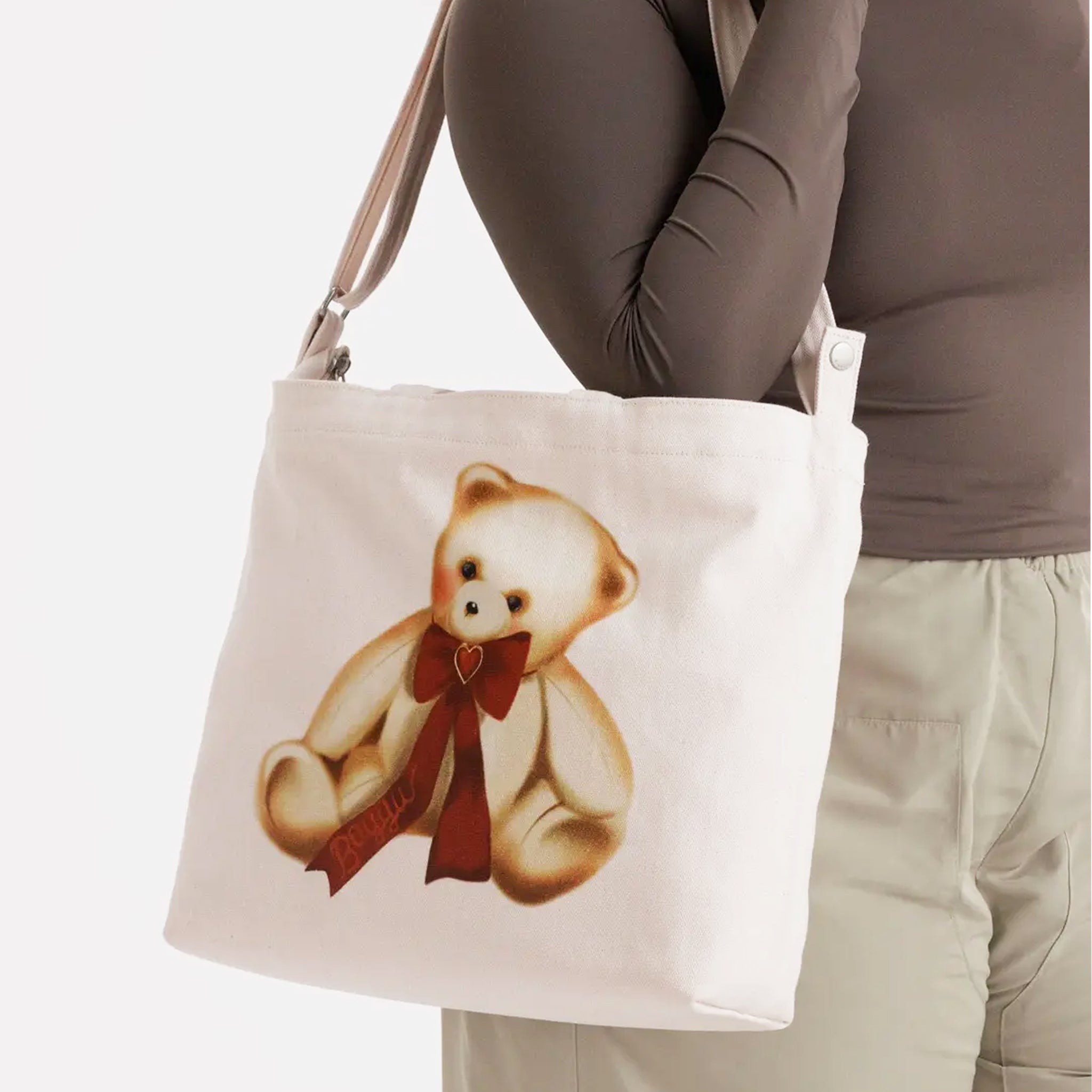 An ivory canvas bag with a shoulder strap and short handle and a teddy bear red bow graphic. 