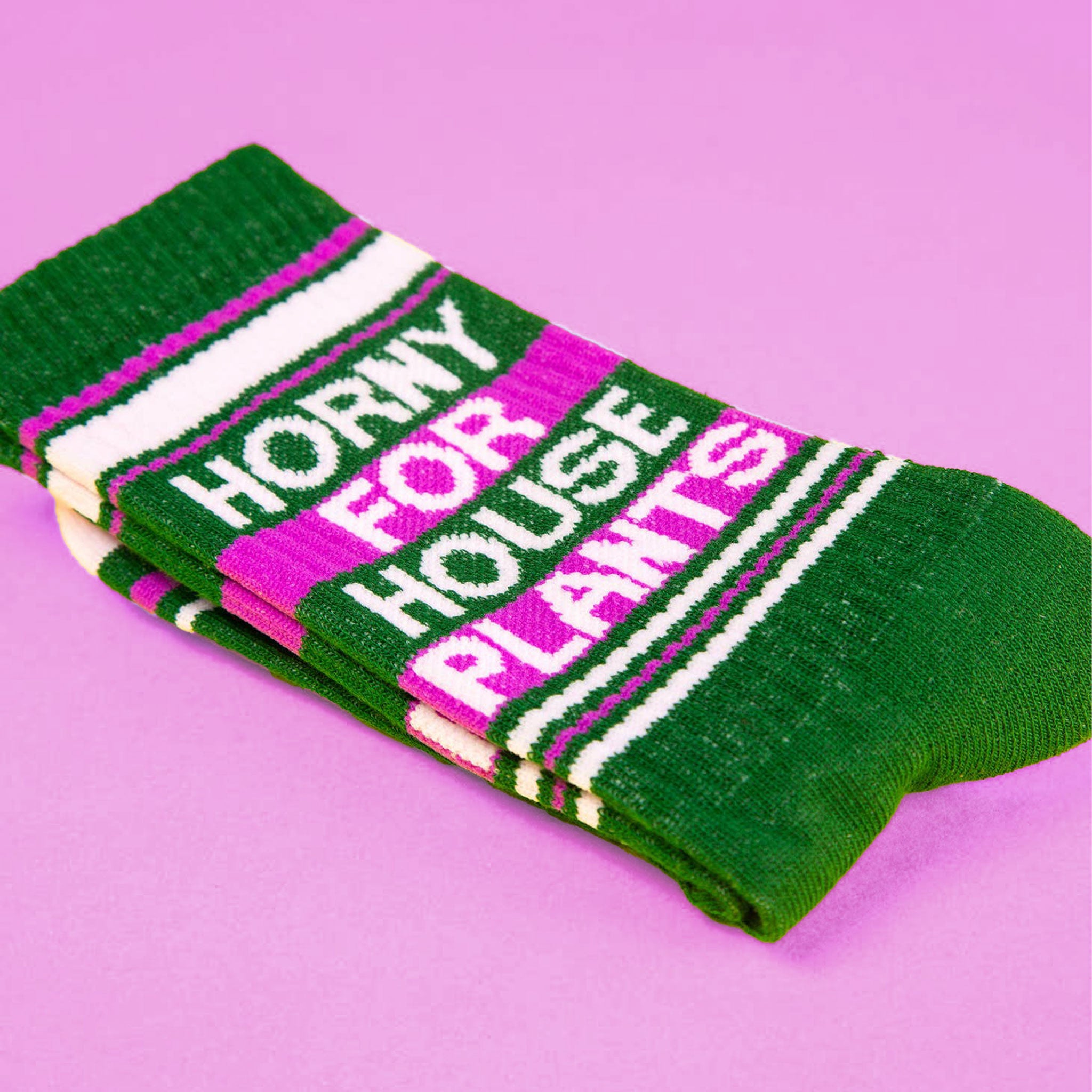 On a purple background is a green and bright pink pair of socks with white text that reads, &quot;Horny For House Plants&quot;