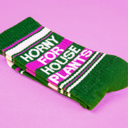 On a purple background is a green and bright pink pair of socks with white text that reads, "Horny For House Plants"