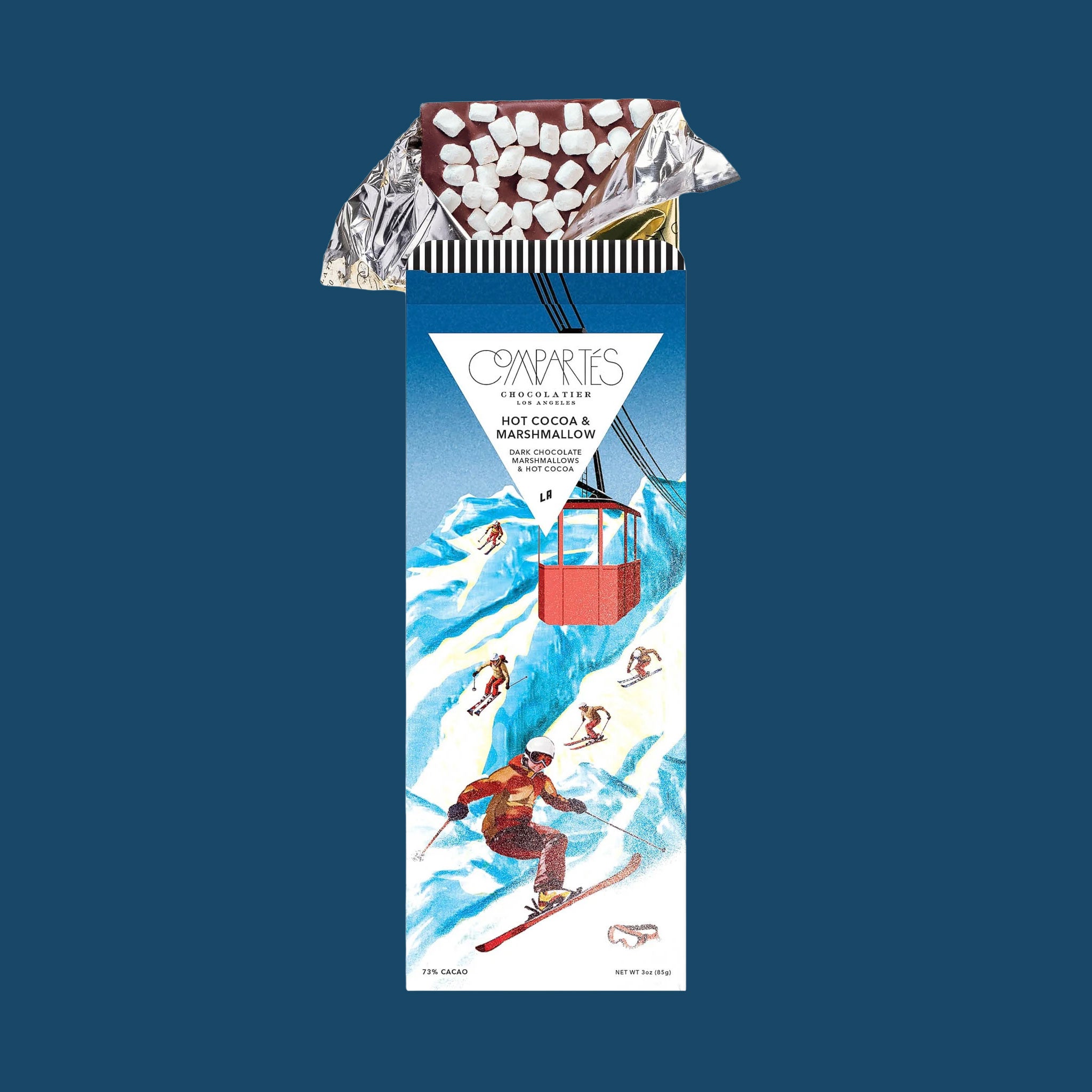 On a dark blue background is a rectangle chocolate bar with packaging that features a mountain and skiers in red outfits along with text at the top that reads, "Compartes Hot Cocoa & Marshmallow Dark Chocolate".