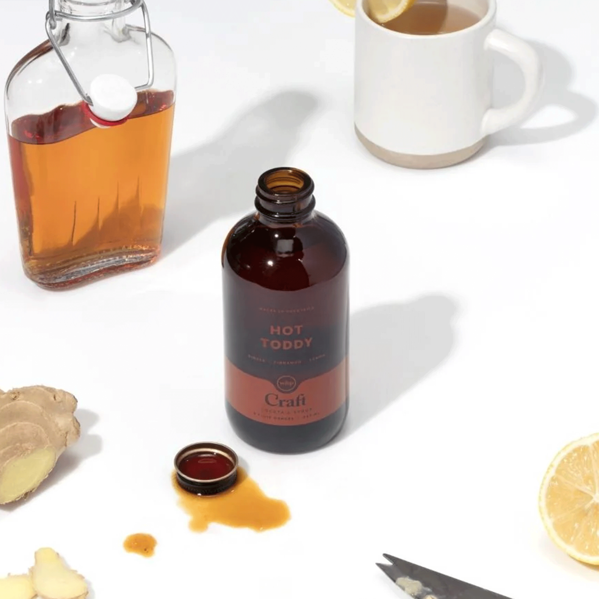 On a white counter is a bottle of hot toddy cocktail syrup in a brown glass bottle with a reddish label. 