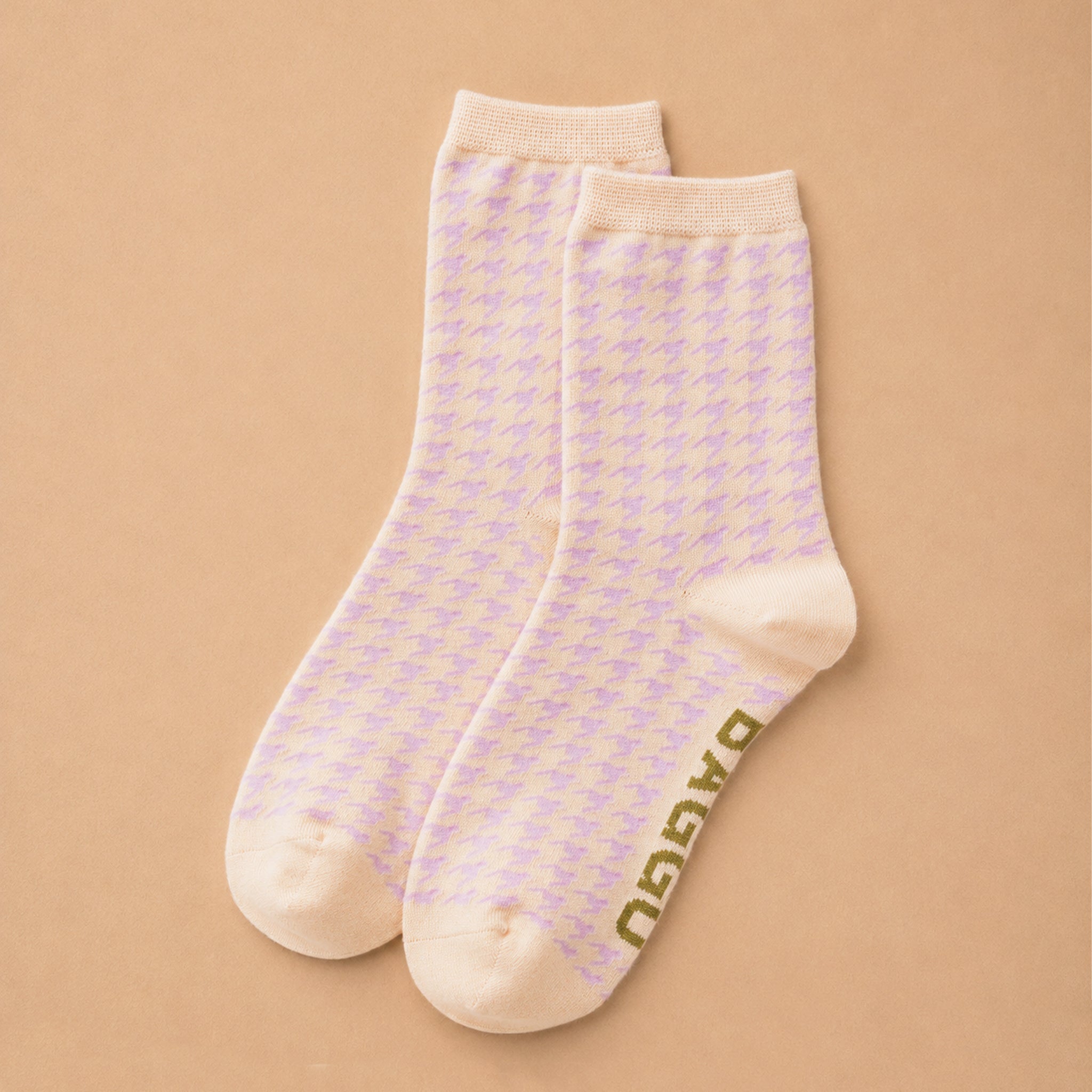 A pair of light cream crew socks with a subtle lavender houndstooth pattern and olive green 'BAGGU' branding printed on the sole, displayed on a warm beige background.