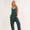 On a neutral background is a model wearing a hunter green tank jumpsuit with big front pockets, a subtle V-neck and a slouchy fit. 