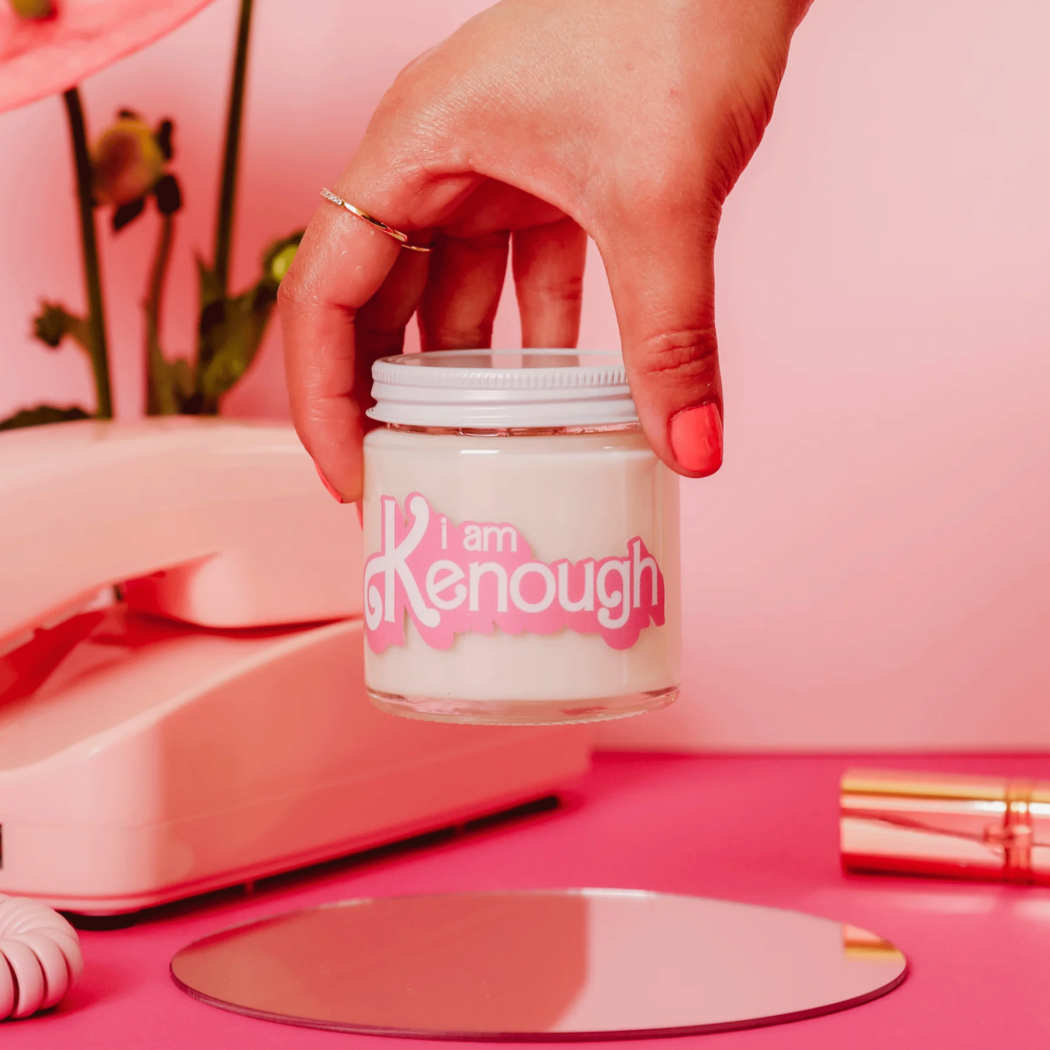 On a pink background is a model holding a glass candle with a white lid and pink text on the front that reads, &quot;I am Kenough&quot;.