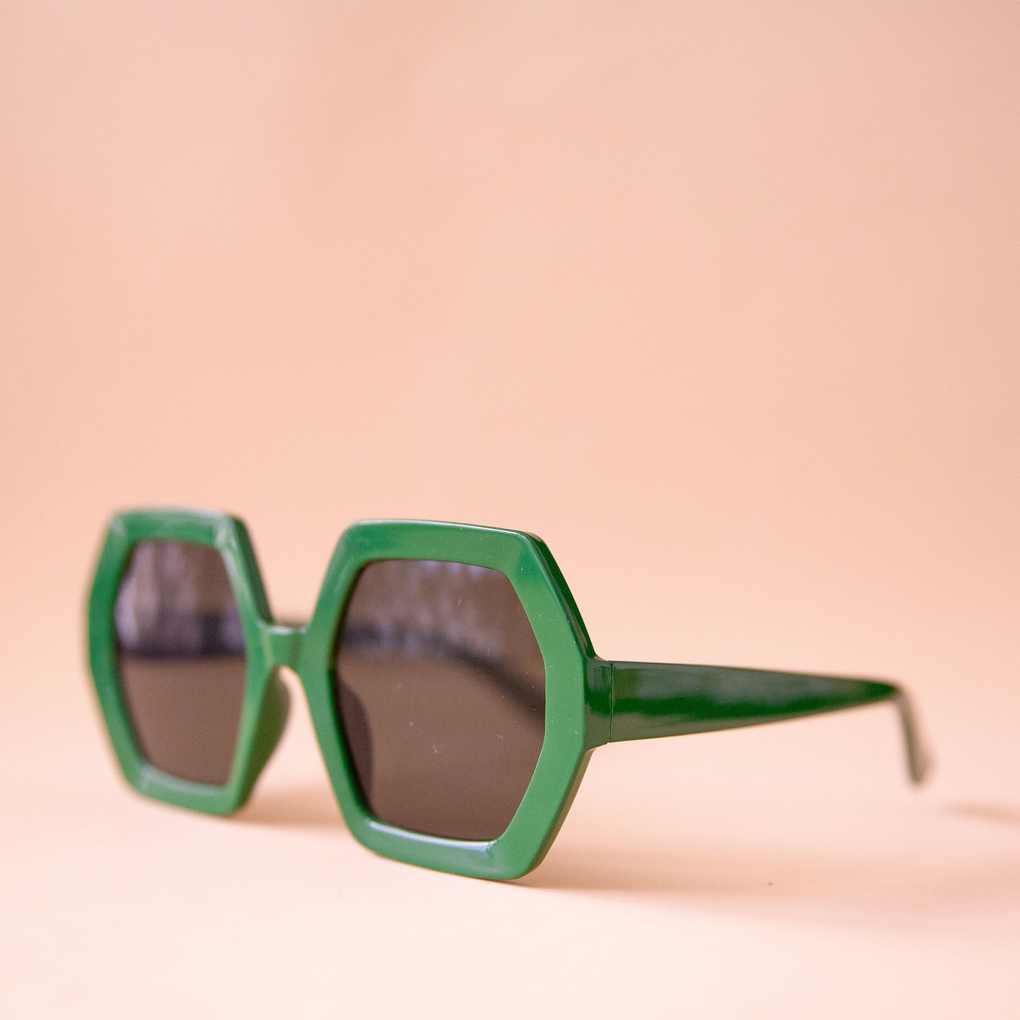 On a peachy background is a pair of hexagon shaped plastic sunglasses in an emerald green color with a dark brown lens.