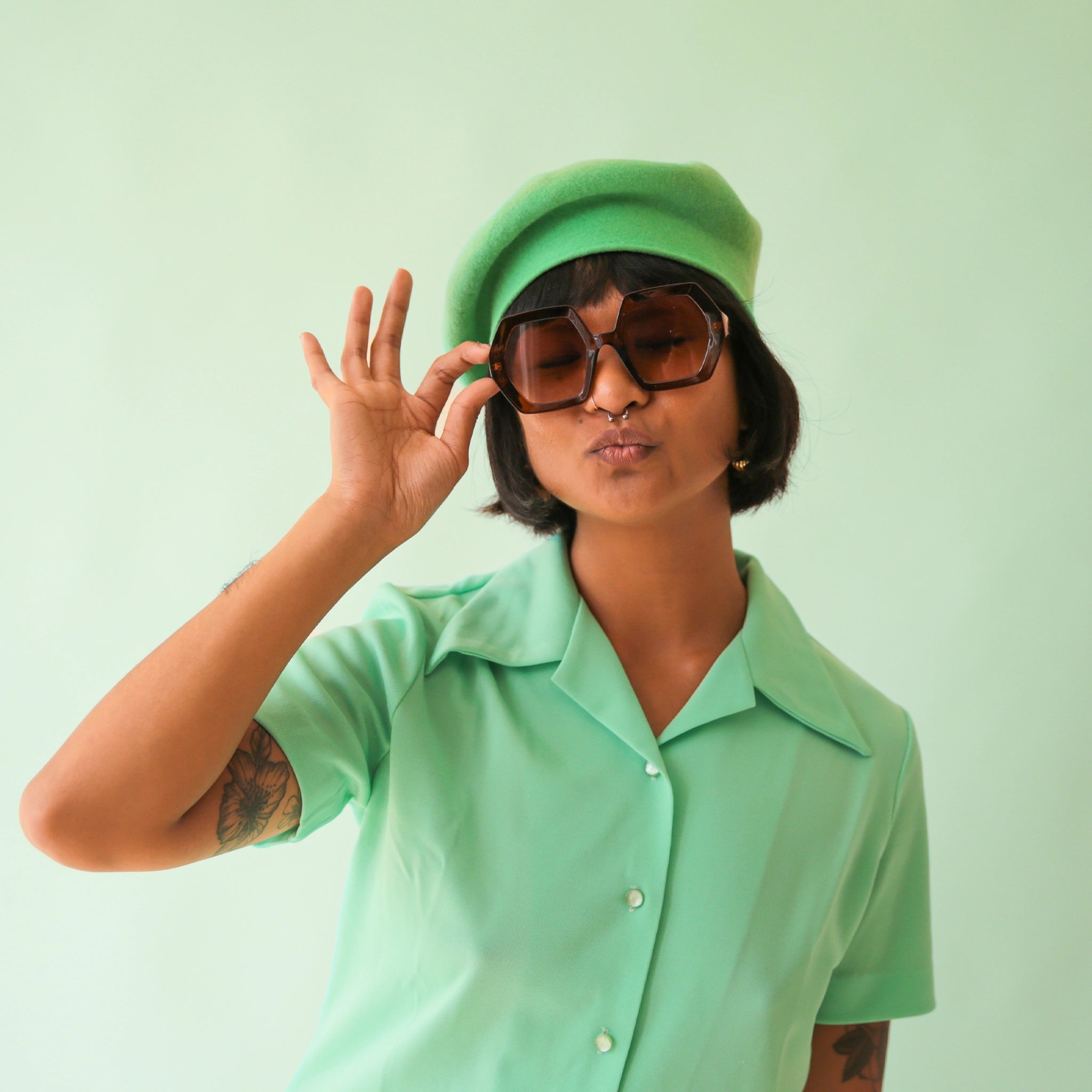 On a green background is modeling wearing a pair of hexagon shaped tortoise sunglasses with brown lenses.
