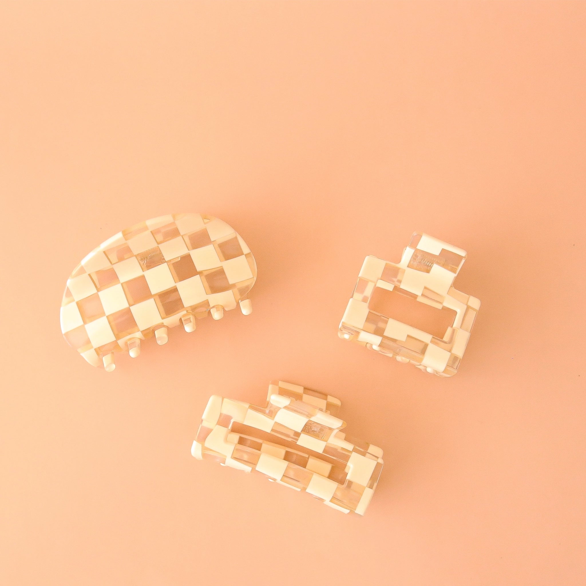 On a peachy background is three different shaped claw clips with a white and clear checker print. One clips is a slightly smaller square shape, the other is a rectangular shape and the third has a rounded edge.