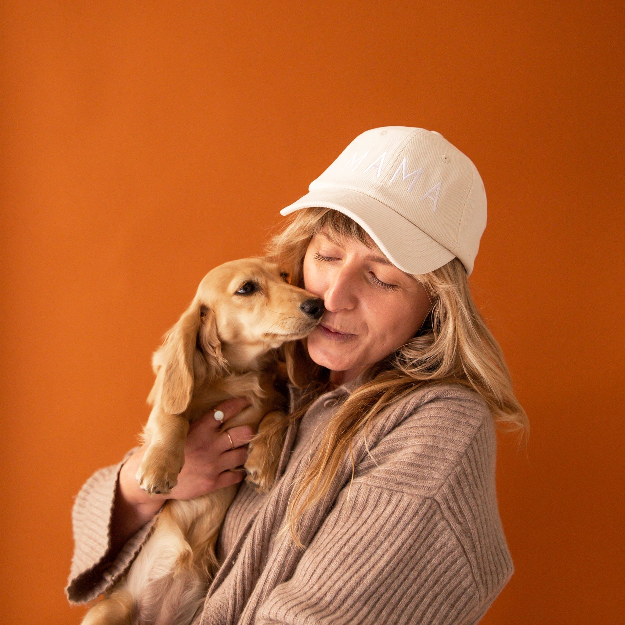 On an orange background is a model holding a dachshund dog and wearing an ivory baseball hat embroidered with white text that reads, &quot;MAMA&quot;.   
