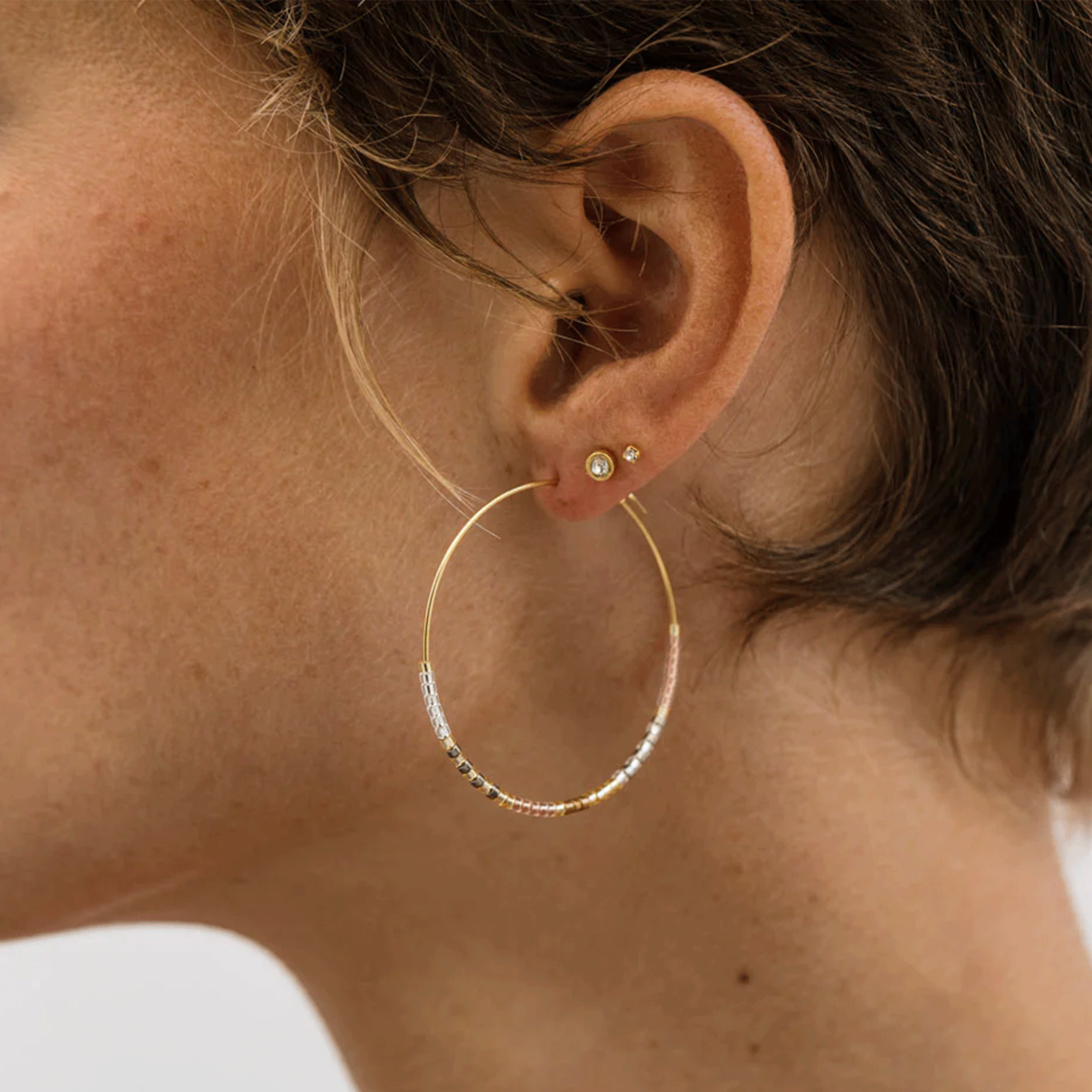 A model wearing a pair of gold thin hoop earrings with neutral colored beads on the bottom half of it. '