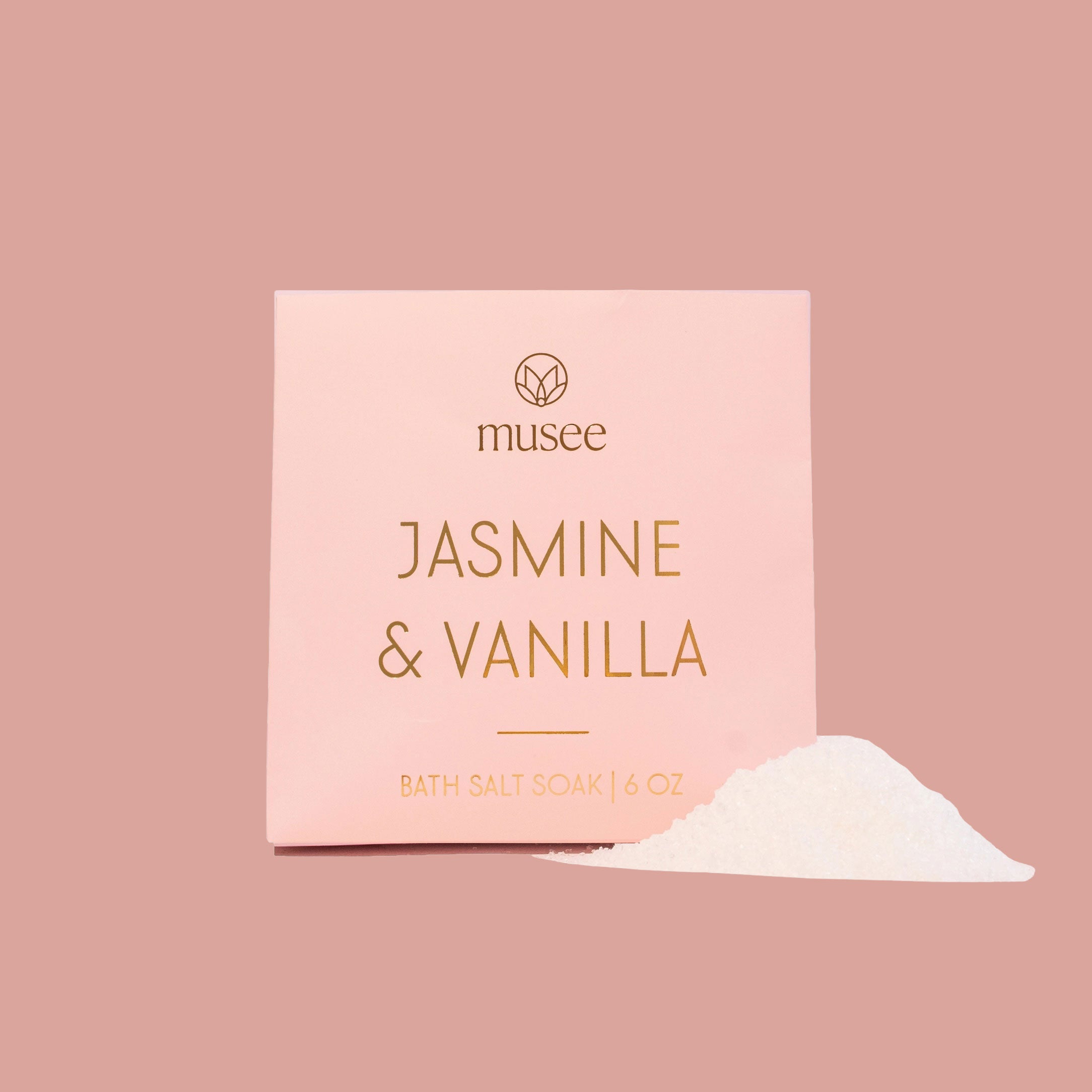 A soft pink Musee bath salt soak package with gold lettering reading “Jasmine & Vanilla” is displayed upright on a dusty rose backdrop. The simple design features clean lines and a minimal aesthetic.
