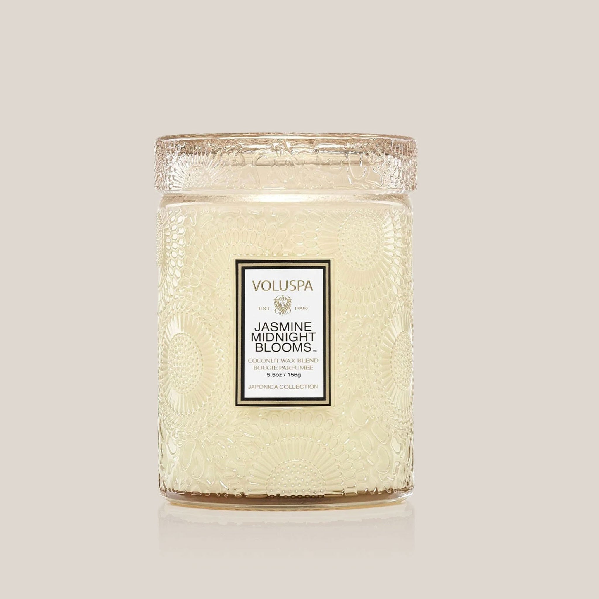An apricot colored decorative glass jar candle with an etched floral pattern in the glass and a white rectangular label in the center that reads, "Jasmine Midnight Blooms".