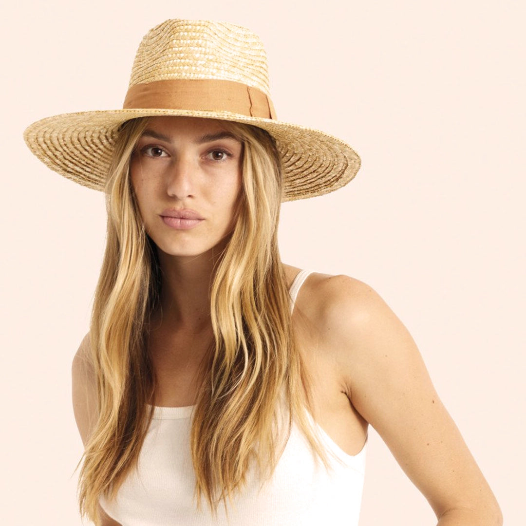 On a cream background is a model wearing a light honey colored straw sun hat with a tan ribbon around the base.