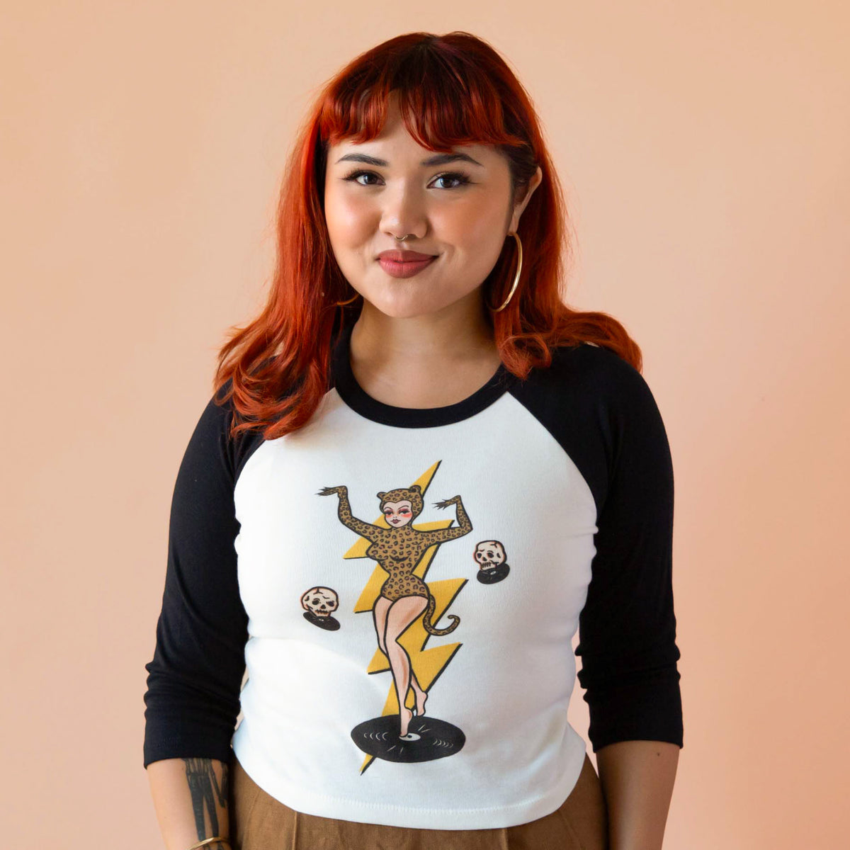 Model with red hair wearing a 3/4 sleeve top, which has a pinup girl in a leopard leotard on it. 