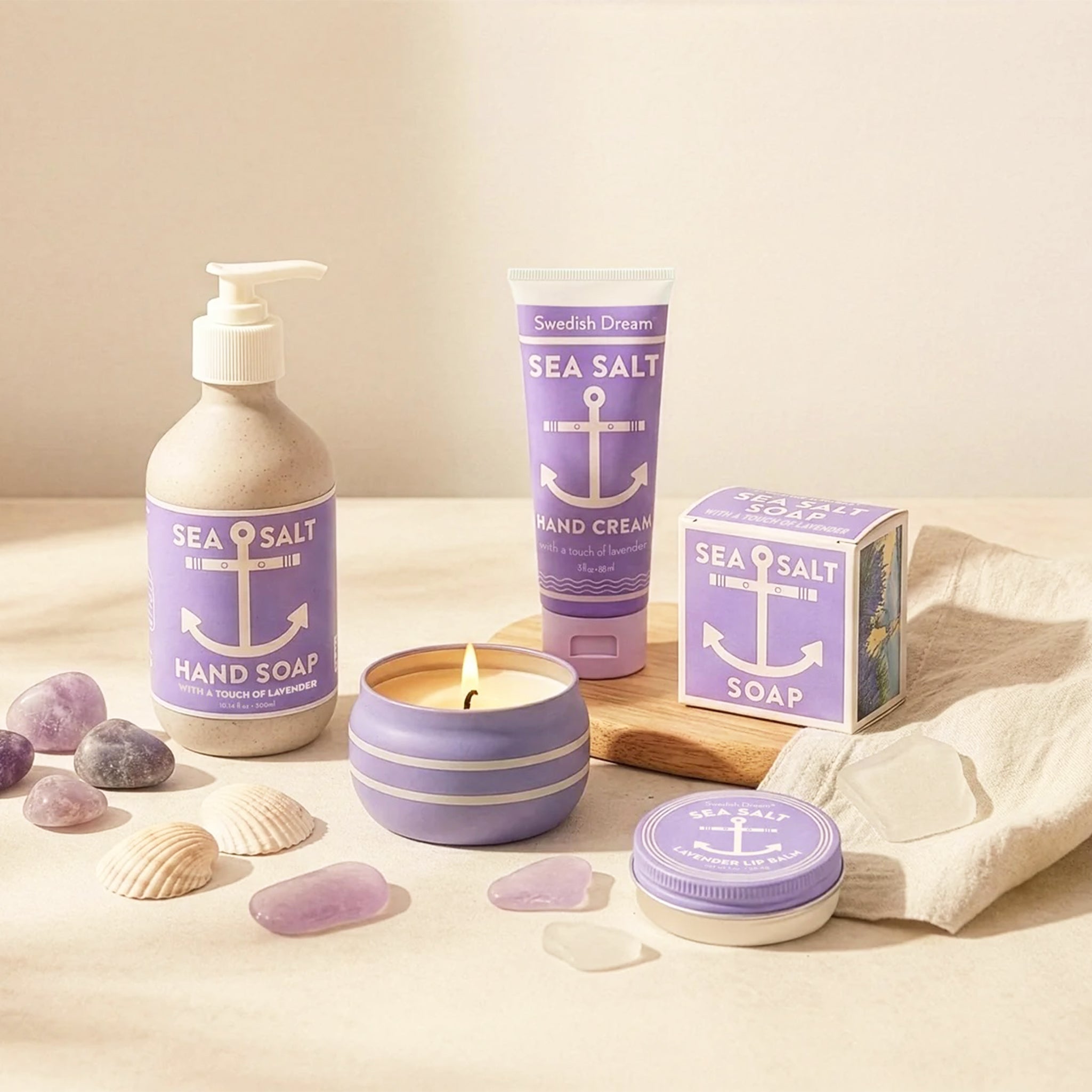 Lavender-themed bath and body products including sea salt hand soap, hand cream, bar soap, lip balm, and a lit candle, styled with seashells and stones on a neutral background.