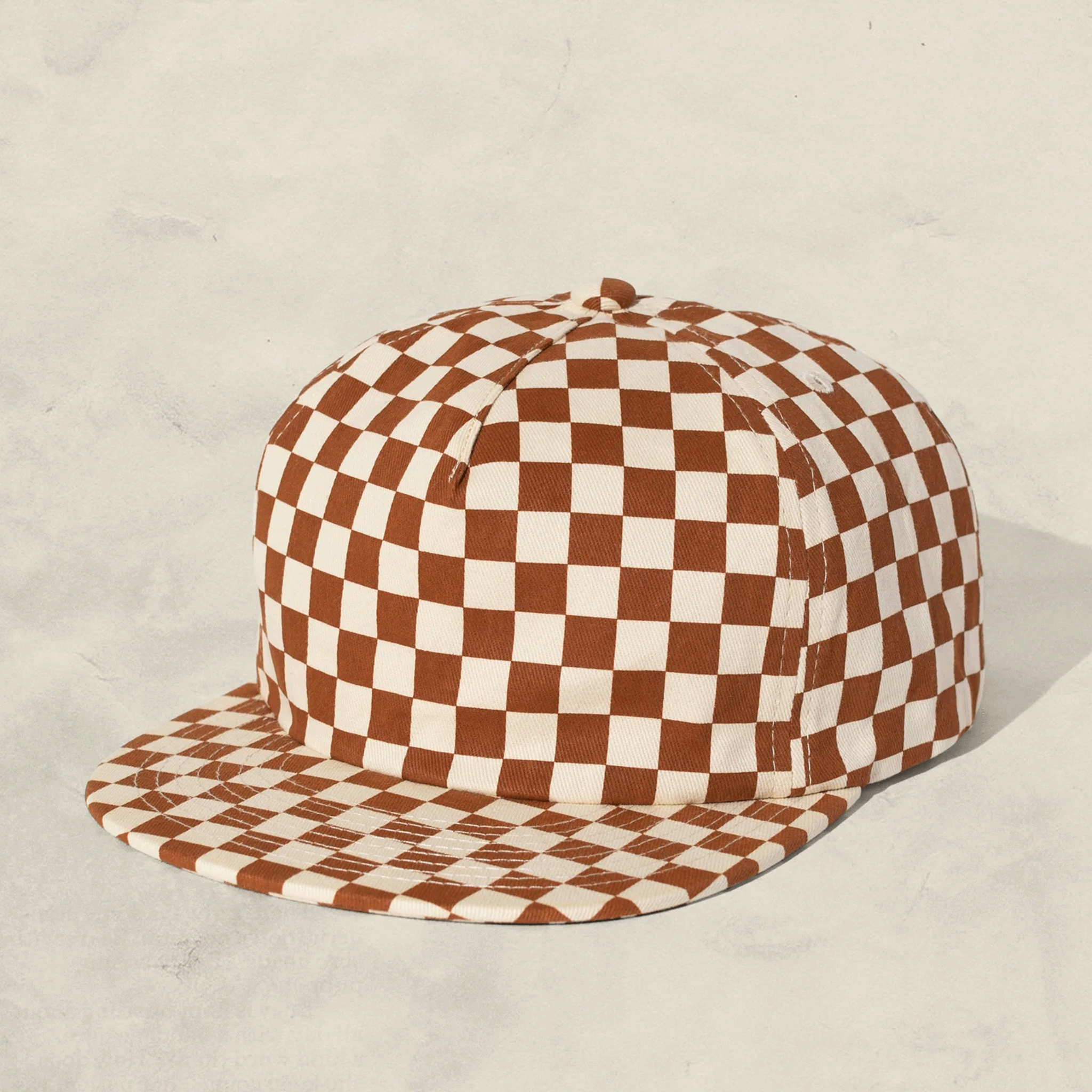 On a grey background is a rust and ivory checker pattern baseball hat for kids. 