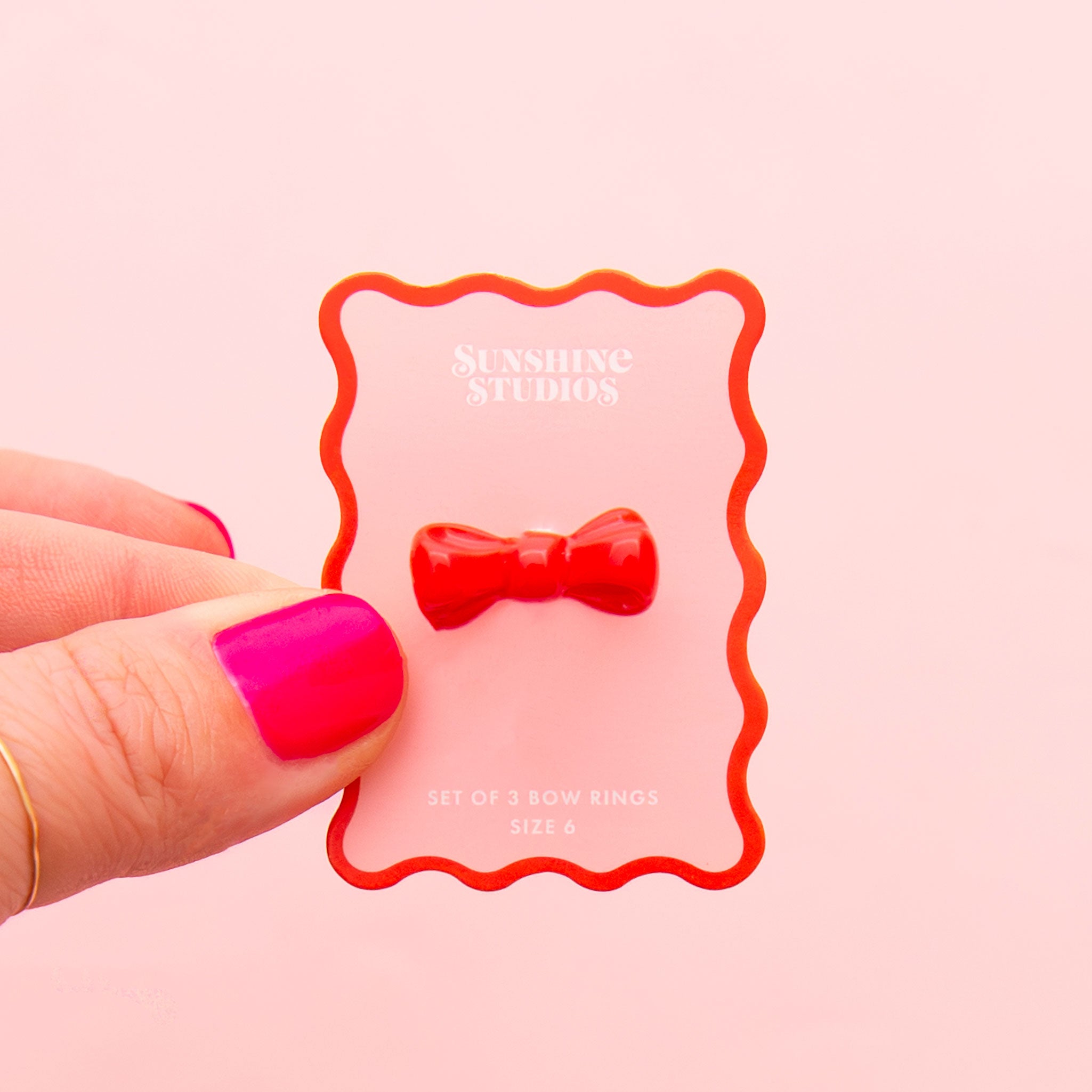 A red bow shaped ring in the middle of the image on a pink backer with red sqiggle border.