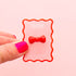 A red bow shaped ring in the middle of the image on a pink backer with red sqiggle border.