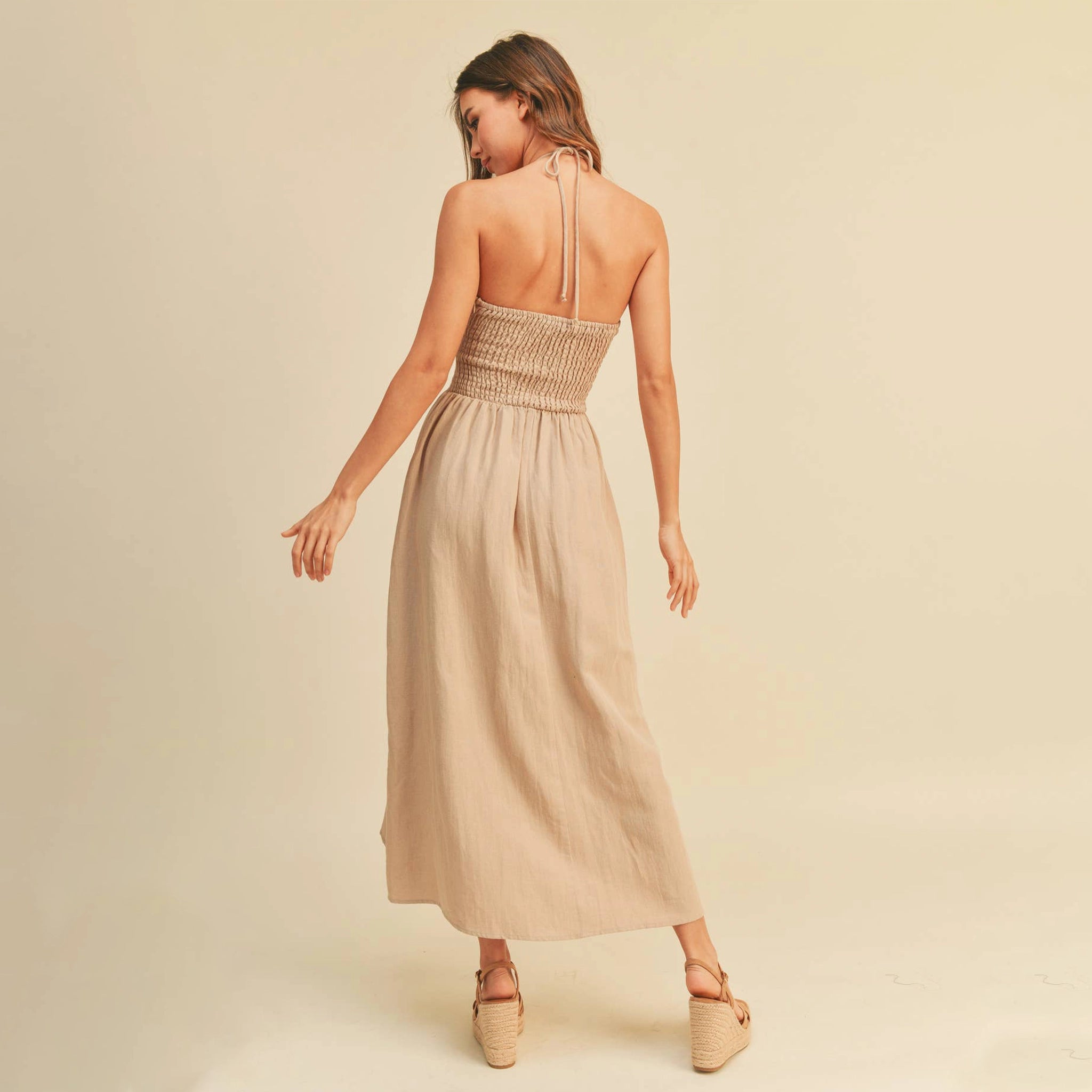 On a tan background is a model wearing the neutral Knotted Front Halter Neck Dress in the shade stone.