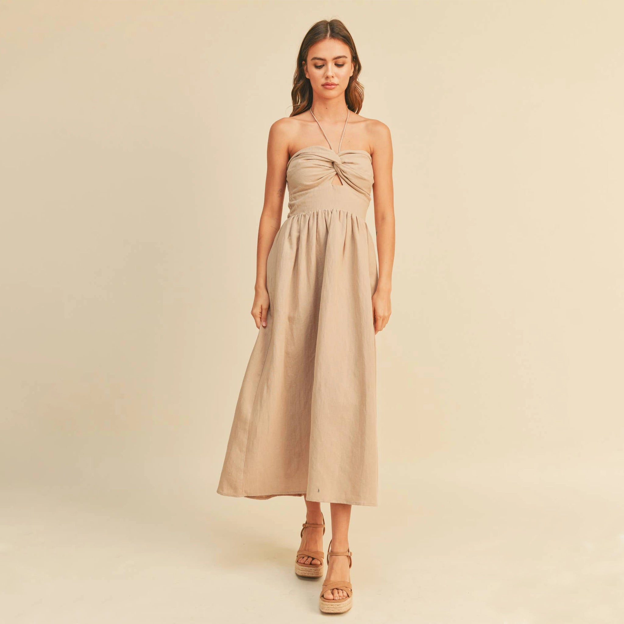 On a tan background is a model wearing the neutral Knotted Front Halter Neck Dress in the shade stone. 