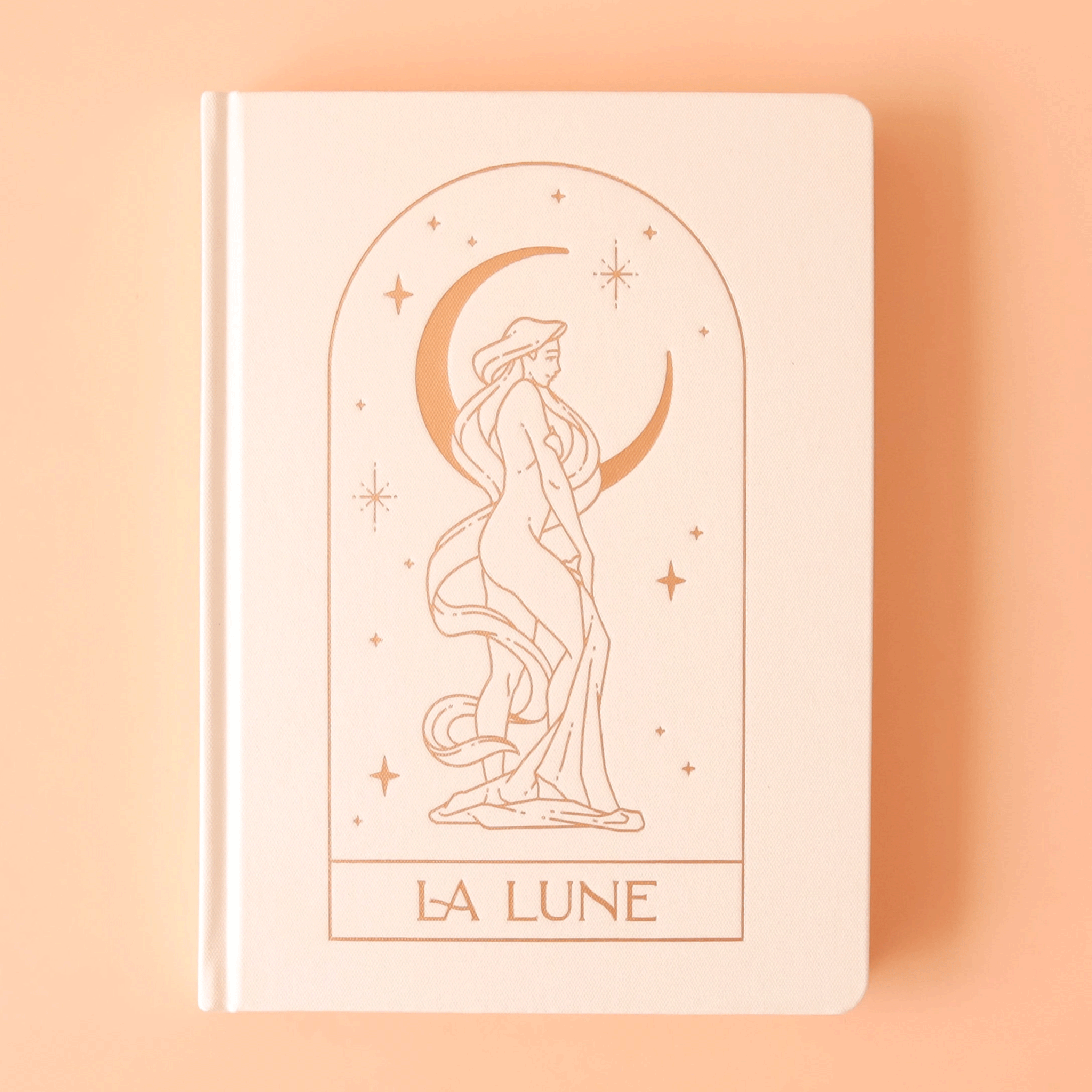 On a light yellow background is a cream hard cover journal with a gold illustration of a person wrapped in cloth in front of a crescent moon and text at the bottom of the arch that reads, &quot;La Lune&quot;.