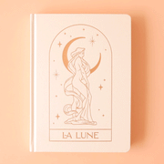 On a light yellow background is a cream hard cover journal with a gold illustration of a person wrapped in cloth in front of a crescent moon and text at the bottom of the arch that reads, "La Lune".