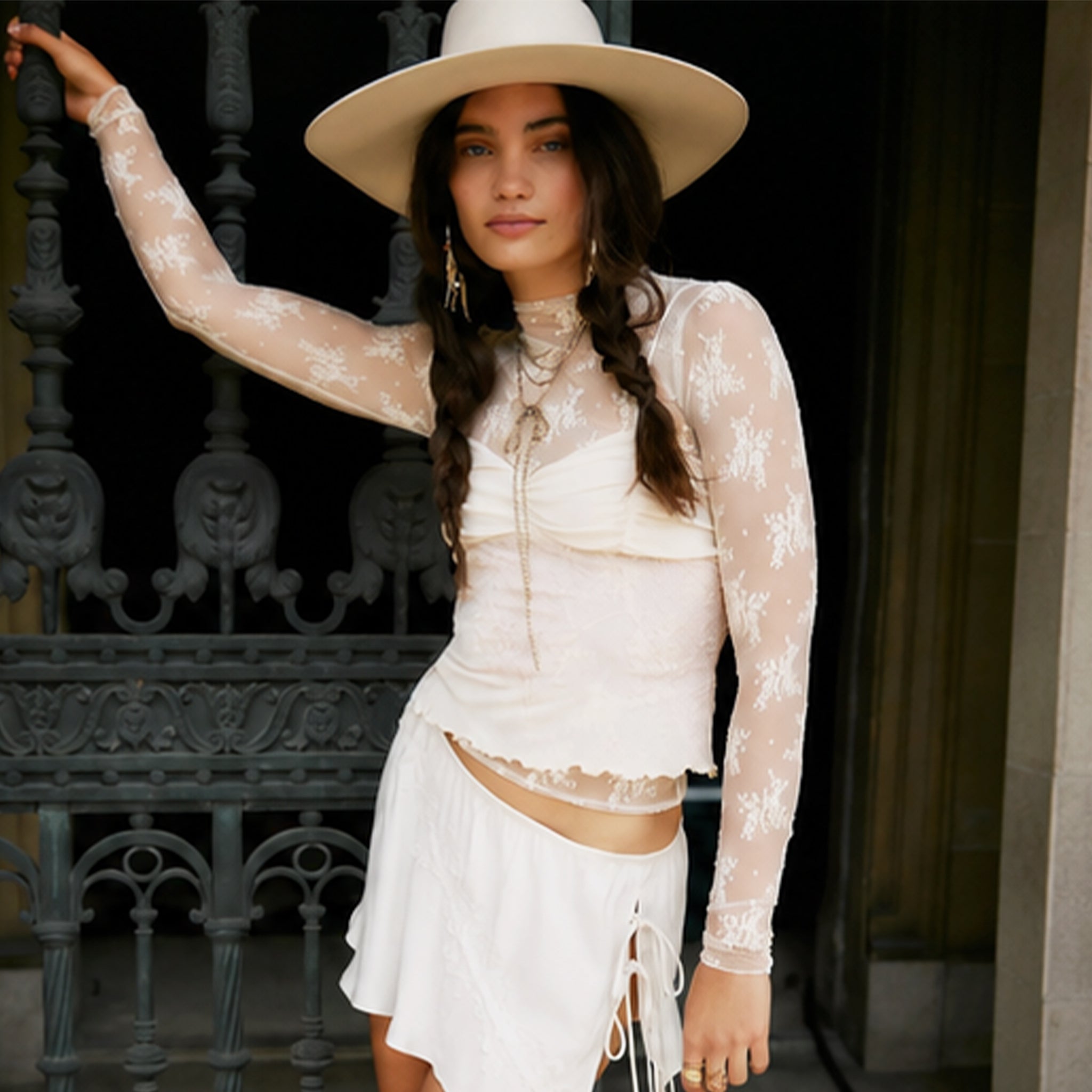 Woman wearing a white lace outfit and hat, standing against a dark background.