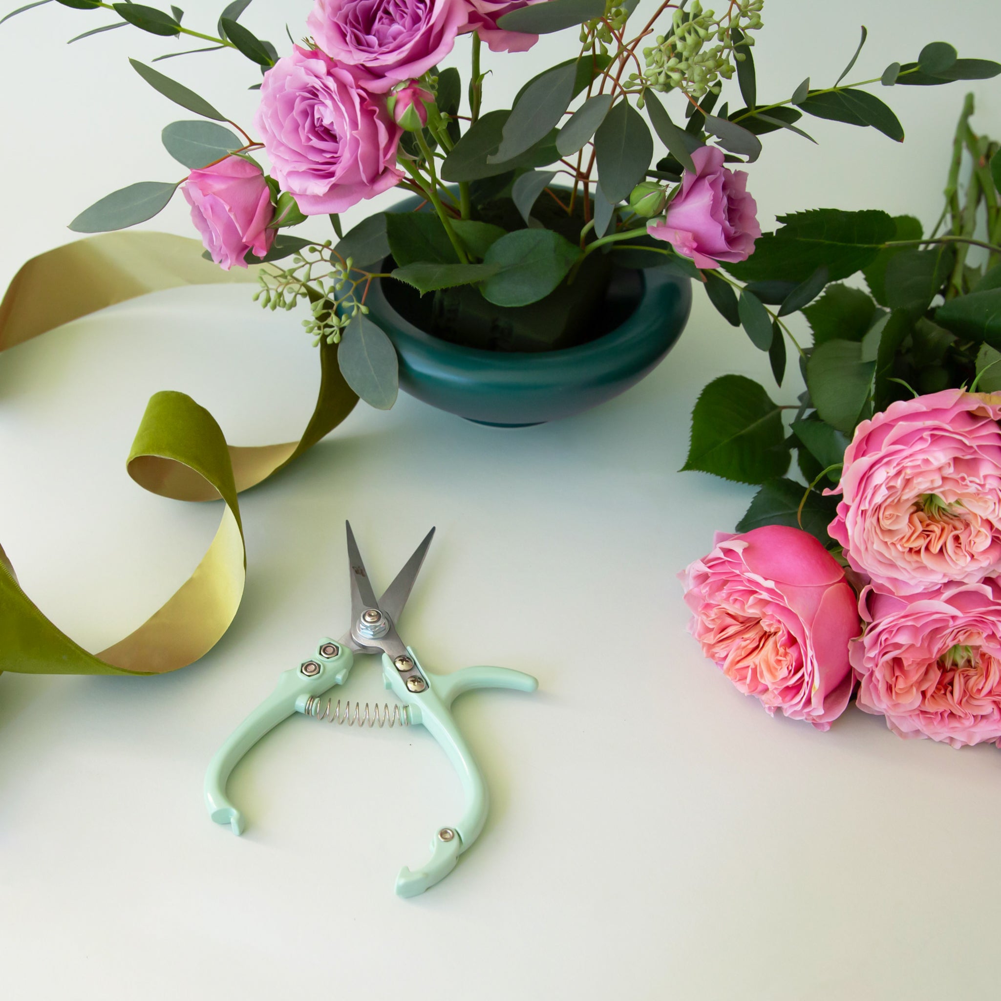 A pair of mint green pruning shears lies in middle of image surrounded by fresh flowers and a dark green bowl with flowers in it.