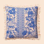 On a cream background is a blue and ivory tropical print pillow with a tassel detail along the edges. 