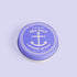 A lavender color lip balm container with an anchor motif.  The lip balm is on a light lavender color background.