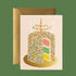 A light pink card with a multicolored cake illustration on the front that has a single candle and a happy birthday sign on top. Also included is a coordinating gold envelope.