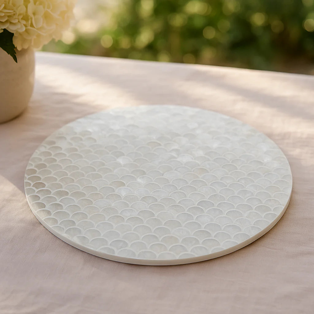 A white capiz lazy susan on a pink tablecloth with green plants in the background.
