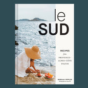 A book cover with a photo of someone cutting fruit on rocks next to the ocean and black text that reads, 'le sud Recipes from Province Alpes-Cote Dazur'. 