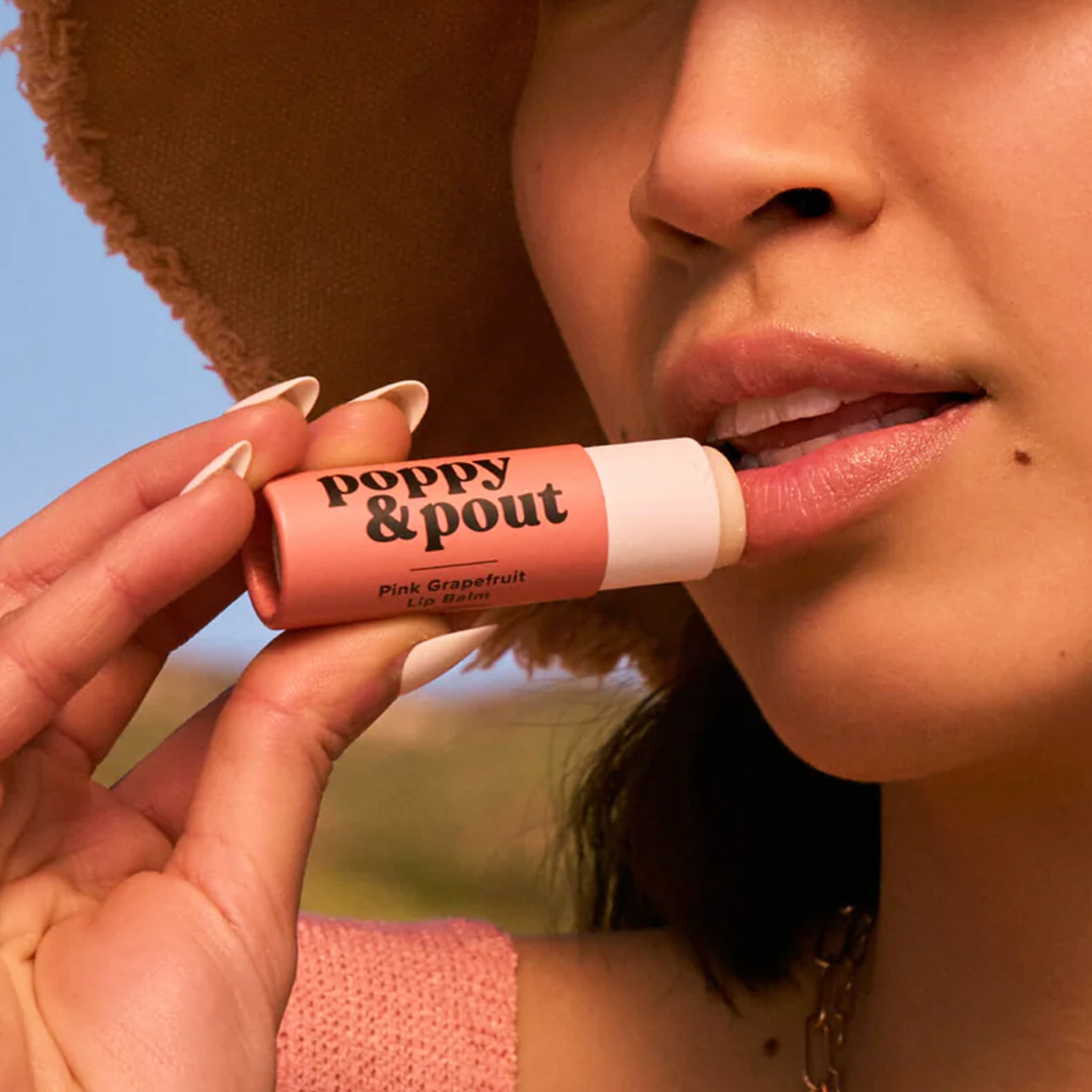 This is a close up picture of a model's mouth. She is holding a round peach tube up to her lips. On the left side of the tube is black text that reads ‘poppy & pout.’