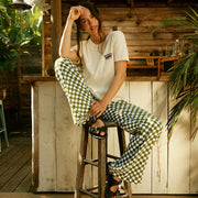 Woman sitting on a wooden stool wearing a white t-shirt and green checkered pants in an outdoor setting.
