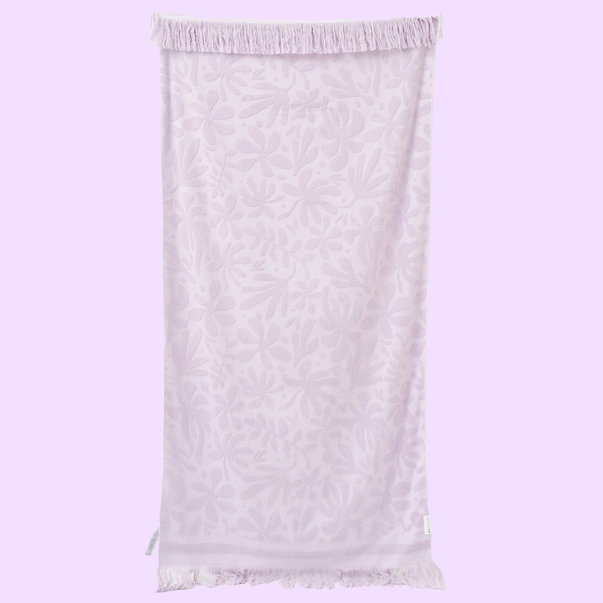 On a purple background is a light purple towel with a subtle floral print and fringe edge. 
