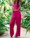 On a green leafy background is a model wearing magenta wide leg cotton pants with an elastic drawstring. 