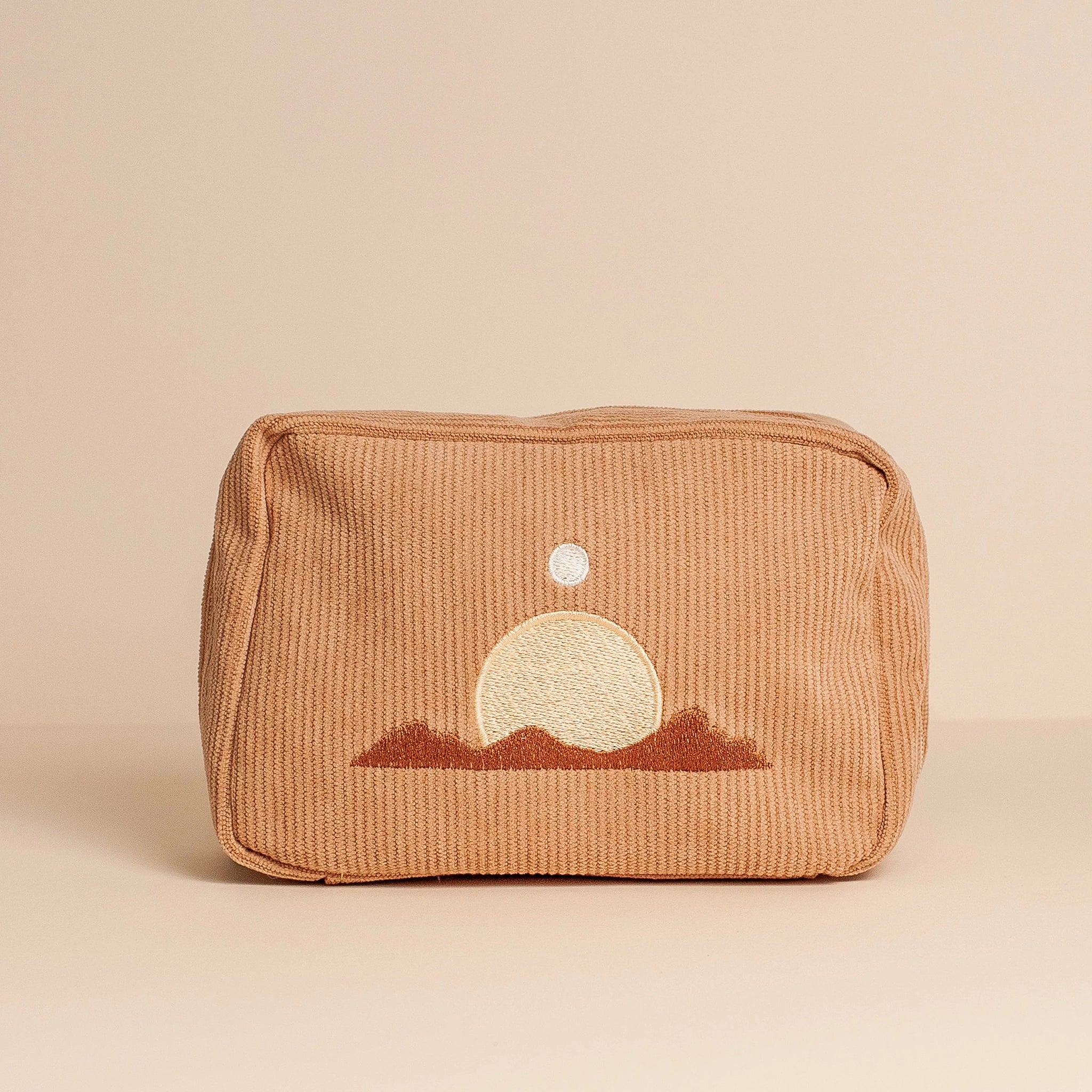 On a tan background is a peachy, tan corduroy makeup bag with a minimalist design of a desert mountain range along with the sun and moon above it. 