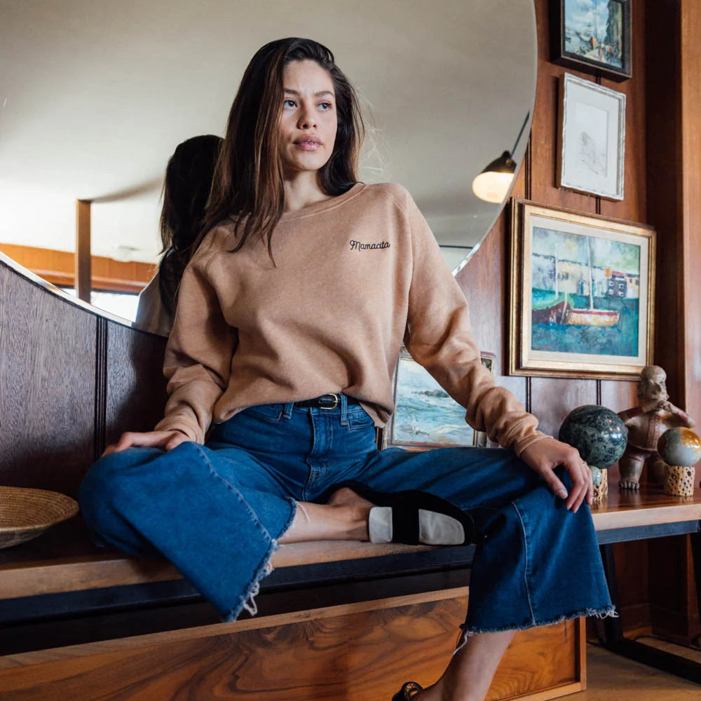 Woman with dark hair seated on a bench wearing a rust colored sweatshirt with &quot;Mamacita&quot; embroidered in black cursive on the front and blue jeans, in front of a round mirror near vintage artwork and decor.