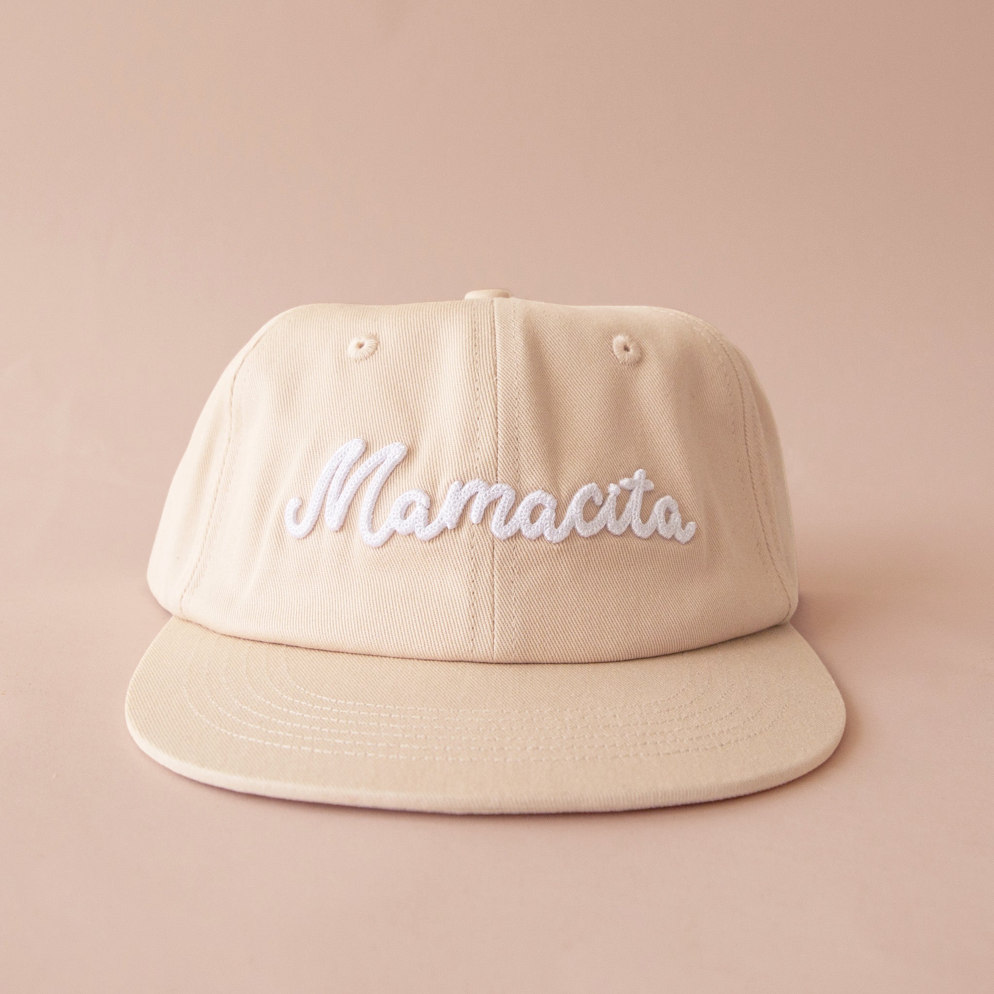 A neutral ivory and white flat brim hat with embroidered text that reads, "Mamacita". 