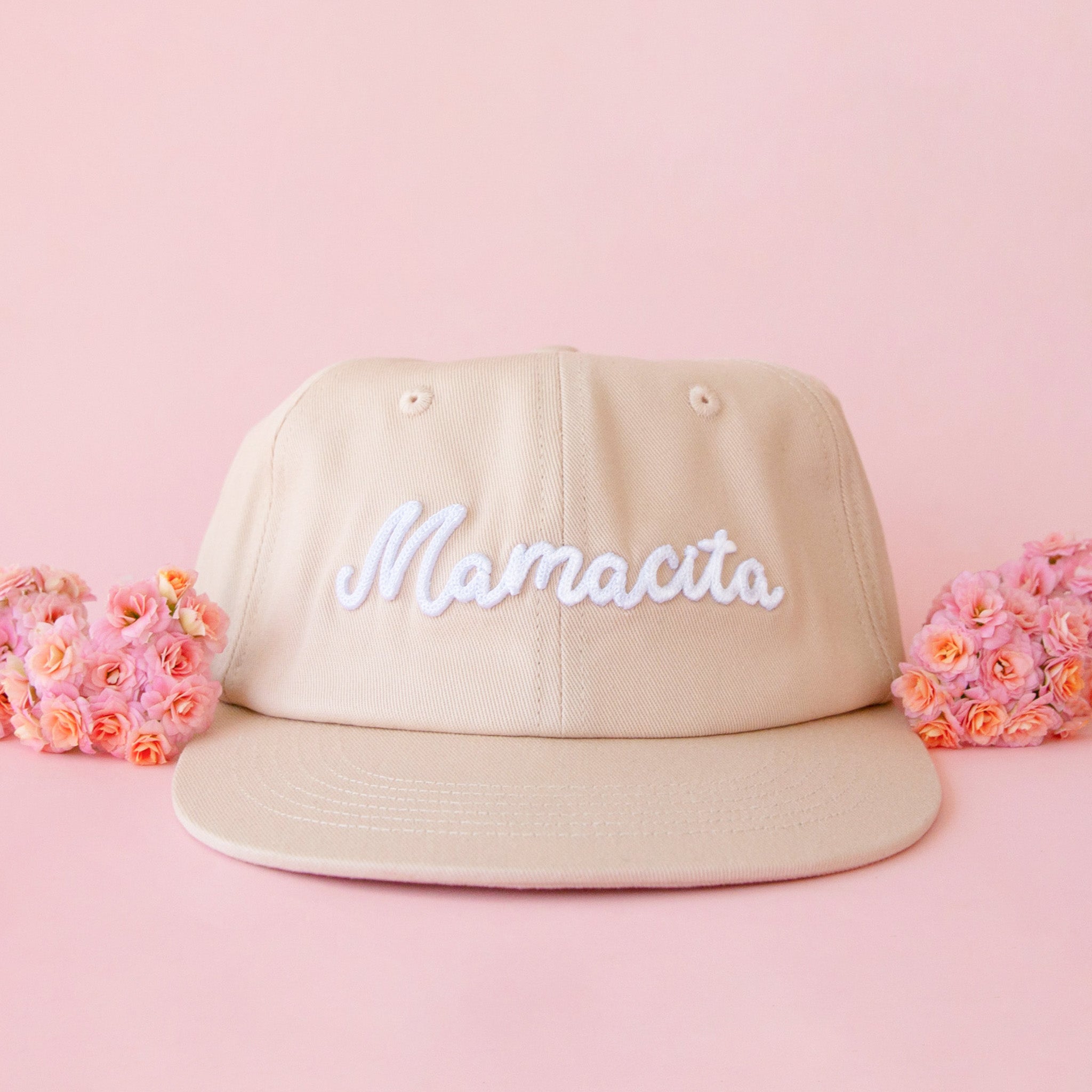 A tan cotton baseball hat that reads "Mamacita" in white embroidery.  There are pink flowers on the sides of the hat sitting next to it.