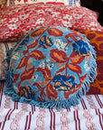 A blue round pillow with red and blue florals with a fringe edge. 